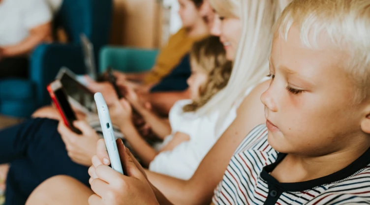 Niño pequeño usando un teléfono móvil mientras adultos y otros menores utilizan dispositivos electrónicos en el mismo espacio.