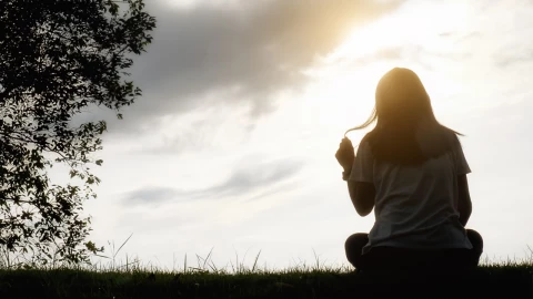 Silueta de una mujer sentada sola sobre el césped al atardecer, con el sol de fondo y cielo parcialmente nublado