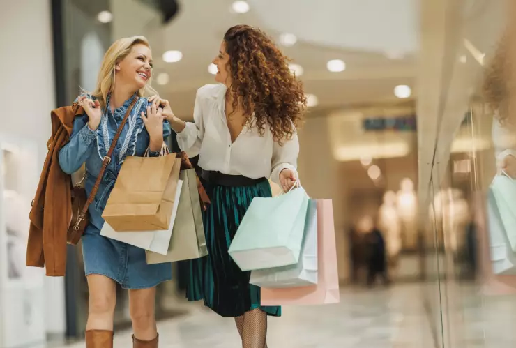 Dos mujeres caminan por el pasillo de un centro comercial cargadas de bolsas y riendo