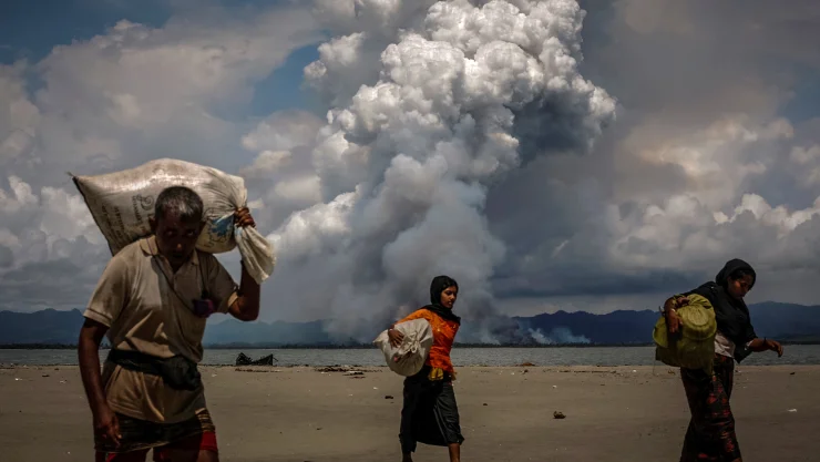 Se ve humo en la frontera con Myanmar cuando los refugiados Rohingya caminan en la orilla después de cruzar la frontera entre Bangladesh y Myanmar en barco a través de la Bahía de Bengala.