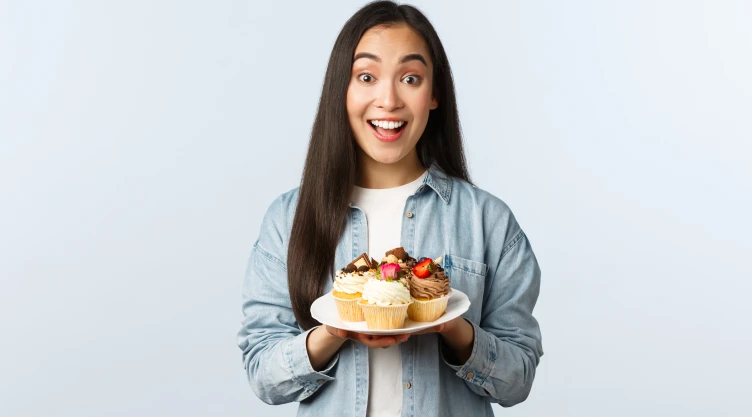 Social distancing lifestyle, covid-19 pandemic, celebrating holidays during coronavirus concept. Excited asian girl holding plate with tasty cupcakes, smiling broadly, invite visit cafe with muffins