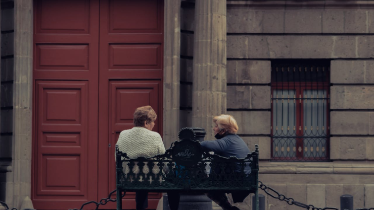 Dos mujeres de la tercera edad se sientan a dialogar frente a un edificio.