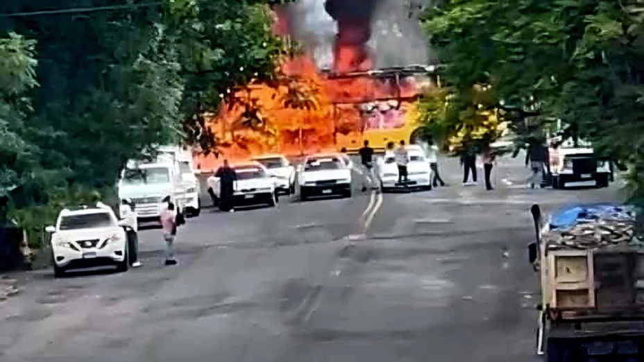 Una escena caótica en Zitácuaro, Michoacán, donde un camión en el fondo de la imagen está envuelto en llamas, creando un resplandor naranja brillante y una densa columna de humo. Varios vehículos, incluyendo coches blancos y camiones, están estacionados a los lados de la carretera, que tiene una línea divisoria amarilla doble en el centro. Numerosas personas están reunidas en la carretera, algunas observando el incendio, mientras otras parecen estar moviéndose. La carretera está flanqueada por una exuberante vegetación y árboles a ambos lados, lo que sugiere un entorno rural o semi-urbano. El cielo está gris y cubierto, y la situación parece tensa y urgente.