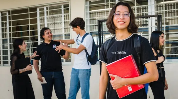 Joven con anteojos sosteniendo un cuaderno rojo frente a más jóvenes felices.