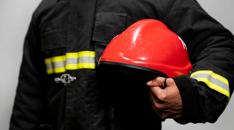 Bombero con uniforme negro y franjas reflejantes sostiene un casco rojo de protección en la mano.