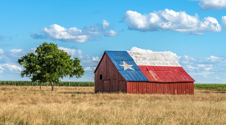 Un pequeño galpón rústico con el techo pintado con la bandera de Texas, en medio de un campo de pueblo