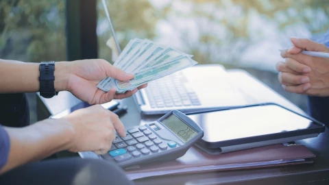 Una persona hace cuentas en una calculadora mientras extiende una mano con dólares