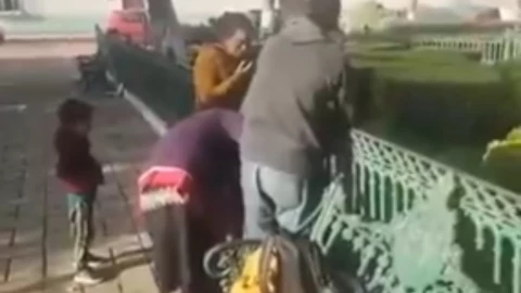 Un plano general muestra a tres adultos y un niño alrededor de un banco de parque de metal verde. El adulto en el extremo izquierdo del grupo de adultos se inclina sobre el respaldo del banco, mientras que otro adulto se inclina sobre el reposabrazos del banco. El tercer adulto está en el extremo derecho del grupo de adultos de pie y mirando hacia abajo en el banco. El niño mira al adulto del extremo izquierdo mientras se inclina sobre el respaldo. La acera está hecha de adoquines de color naranja rojizo. Detrás del banco hay un césped y árboles. En el césped de la derecha hay una zona de césped vallada. En la parte superior de la imagen hay más casas con tejados blancos y naranjas.