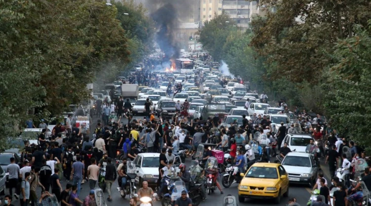 Multitud de manifestantes ocupa una avenida llena de autos detenidos mientras columnas de humo se elevan al fondo, en medio de disturbios durante protestas en Irán.