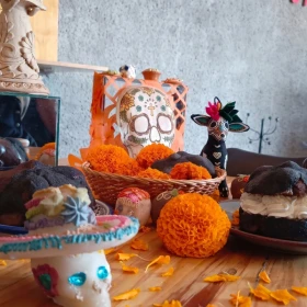 Mesa de madera decorada con piezas de pan de muerto negro, una rellena de crema chantilly blanca y otra con cubierta de chocolate, rodeadas de flores de cempasúchil, calaveritas de azúcar y una taza de barro.