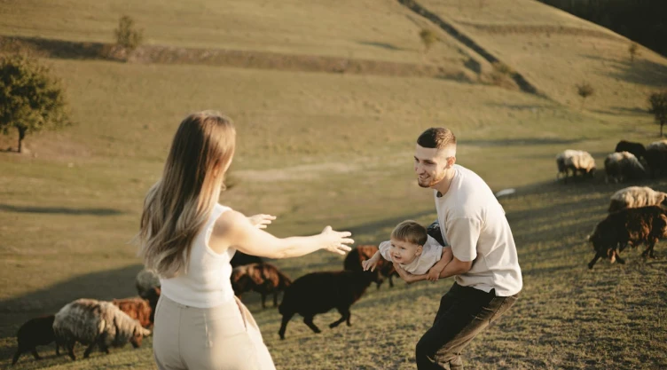 Mujer con los brazos abiertos, juega con su bebé y su esposo vestido de blanco en el campo.