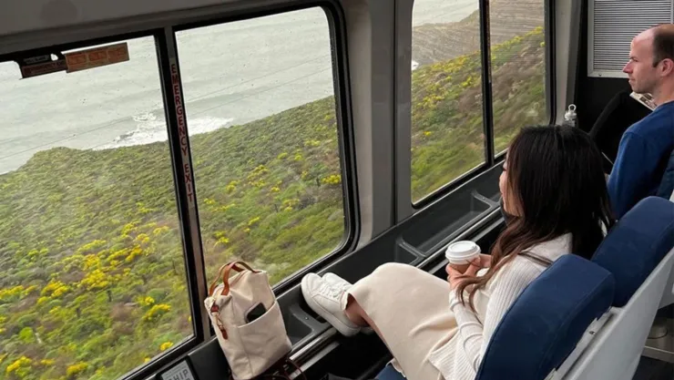 Dos pasajeros sentados con un café frente a las grandes ventanas en las que se ve el paisaje.