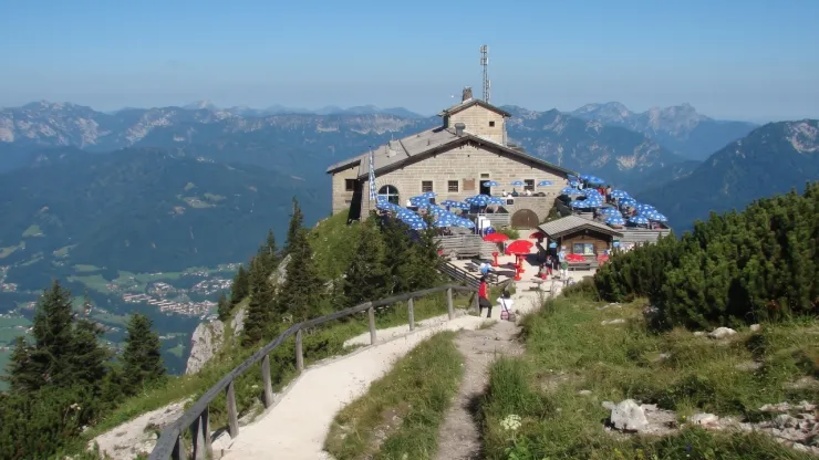 El Nido del Águila como destino turístico Vista diurna del edificio Kehlsteinhaus, conocido como El Nido del Águila, en la cima de una montaña soleada con turistas y sombrillas azules y rojas en su terraza.en los Alpes de Baviera antes del incendio