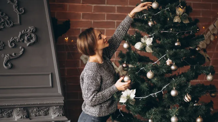 una mujer colocando adornos del árbol de navidad: esferas doradas y un moño plateado acompañados de luces