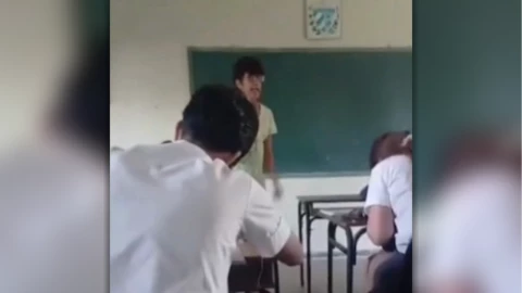 Estudiante habla frente a su profesora dentro de un salón de clases en Cuba mientras otros alumnos permanecen sentados en sus pupitres.