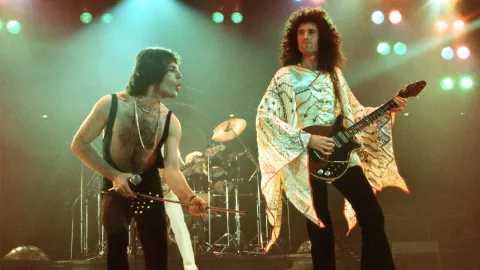 Freddie Mercury cantando vestido de negro y Brian May tocando la guitarra con playera blanca larga cuando eran jóvenes