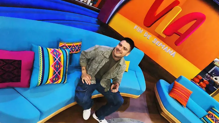Conductor de televisión saludando y sentado en una sala color azul dentro de un foro de televisión con una pantalla gigante detrás