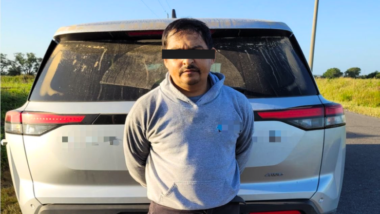 Sujeto mexicano buscado por abusar de una niña de 12 años está arrestado y con antifaz en los ojos; porta una sudadera gris; está parado frente a la cajuela cerrada de una camioneta.