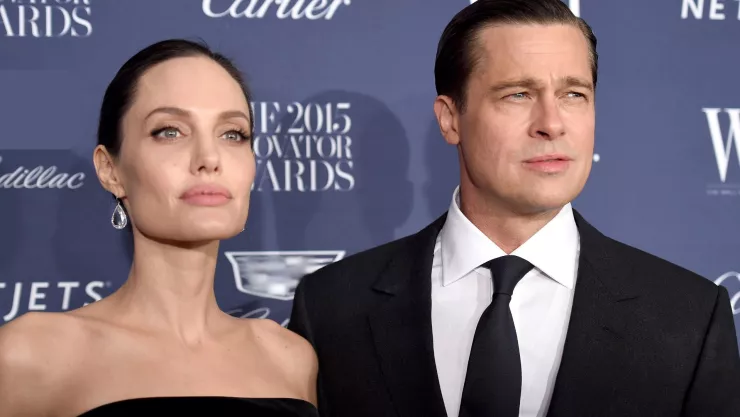 Angelina Jolie y Brad Pitt se encuentran en una sesión de fotos, ella lleva vestido negro, cabello recogido y aretes de diamantes, él lleva traje de vestir negro