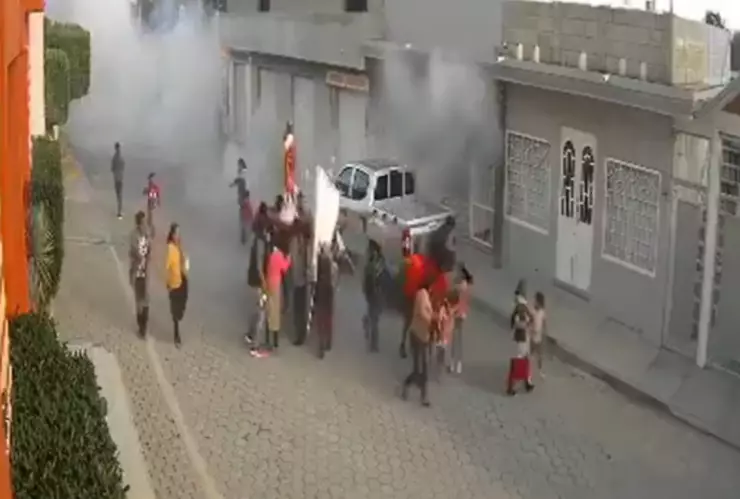 Una vista aérea muestra una concurrida calle de la ciudad con personas y vehículos. En el lado izquierdo, un edificio alto se eleva sobre el resto, dominando el horizonte. En medio del humo blanco tras estallido de pirotecnia.