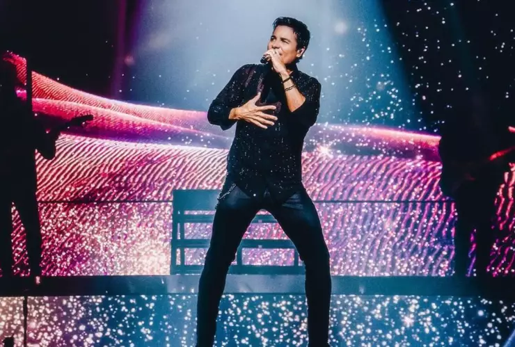 Chayanne con un micrófono cantando en su tour “bailemos otra vez”, vestido de negro con una camisa con brillos
