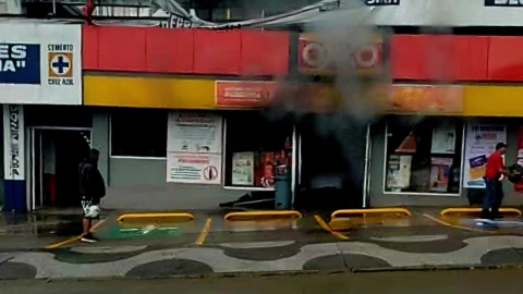Un hombre con pantalones cortos de atletismo y una camisa de manga corta negra está de pie en la calle mojada frente a un edificio con una franja roja sobre una franja amarilla. Hay humo saliendo de la parte superior del edificio, cerca de un cartel de Sika. Un letrero de “CEMENTO CRUZ AZUL” es visible a la izquierda. Un hombre con una camisa roja está de pie a la derecha, sosteniendo lo que parece ser una manguera o un cable. El cielo está nublado y los cables aéreos son visibles en la parte superior.