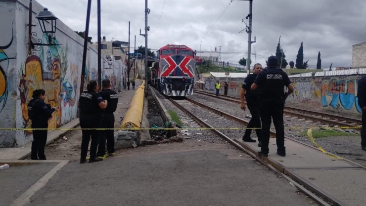 Un tren de carga rojo y blanco con un gran símbolo “X” rojo en la parte delantera está detenido en las vías del tren, que se extienden en la distancia. Varios agentes de policía municipal, están de pie cerca de las vías, y una cinta amarilla de “POLICÍA MUNICIPAL” en Querétaro.