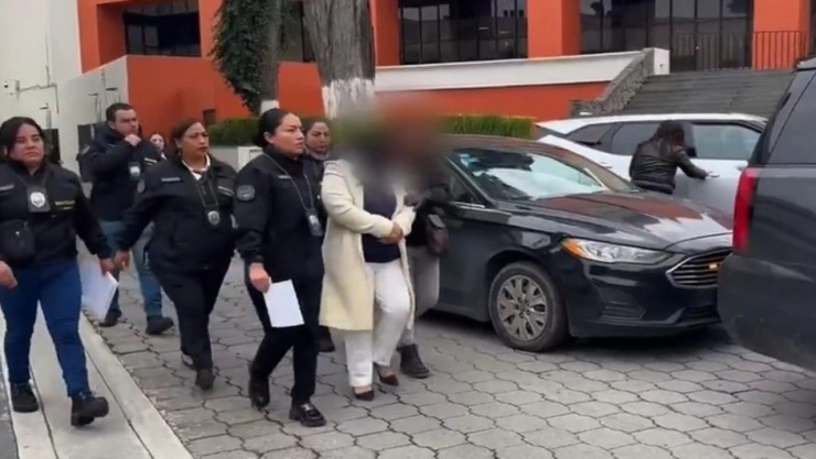 Una mujer de mediana edad, con el cabello recogido y vestida con un traje oscuro, está sentada en un gran sillón de madera tallada con reposabrazos, detrás de un escritorio oscuro. Su rostro está cubierto por una barra negra horizontal para preservar su anonimato. A su izquierda, sobre el escritorio, hay una pequeña bandera de México. El fondo es una pared con azulejos texturizados en tonos claros.