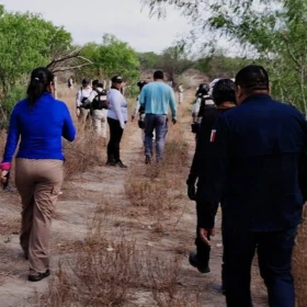Fotografía de una brigada de búsqueda en un terreno árido de Tamaulipas. Se observa a un grupo de personas, integrantes del colectivo ‘Amor por los Desaparecidos’ y elementos de seguridad, caminando de espaldas por un sendero flanqueado por maleza seca y arbustos verdes. Al frente, una mujer con playera azul y pantalón café encabeza el grupo, mientras que otros visten uniformes tácticos o ropa de campo. El ambiente es soleado y el terreno es de tierra suelta, típico de las zonas de búsqueda de restos en el norte de México.