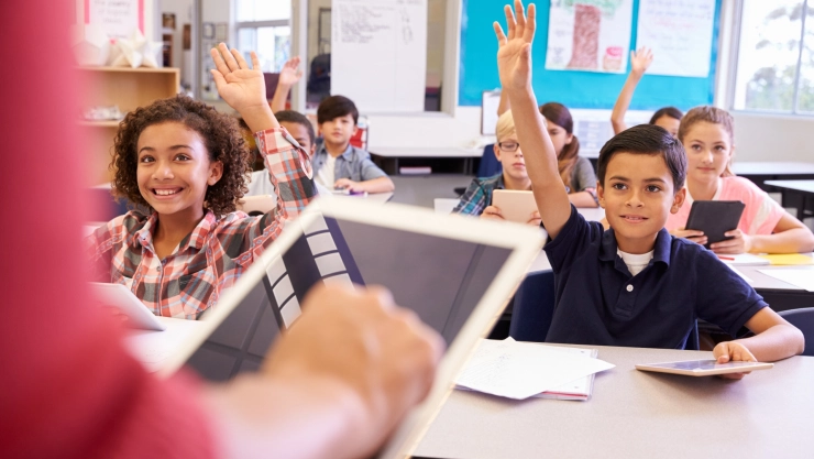 Un grupo de niños levantan la mano ante la maestra en un aula.