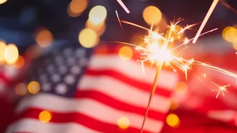 Un pequeño juego pirotécnico adelante de una bandera de Estados Unidos.