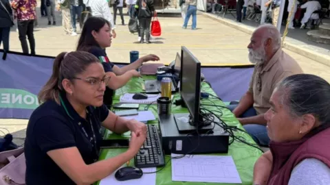 Mujer de playera negra frente a computadora hablando con mujer de chaleco lila.