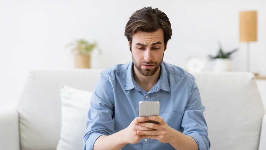 Frustrated Man Using Cellphone Texting Sitting On Sofa At Home