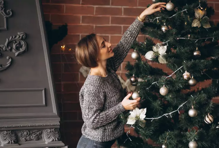 una mujer colocando adornos del árbol de navidad: esferas doradas y un moño plateado acompañados de luces