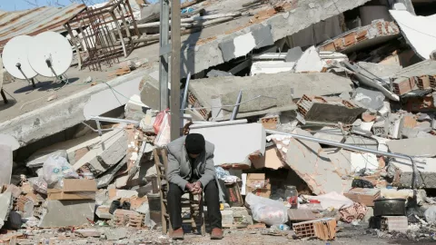 Hombre sentado con la cabeza inclinada, detrás de él solo hay escombros de edificios colapsados
