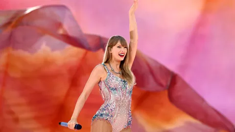 Taylor Swift posando con una mano levantada, sonriendo con labios rojos y su vestimenta de brillos para su concierto del ERAS Tour
