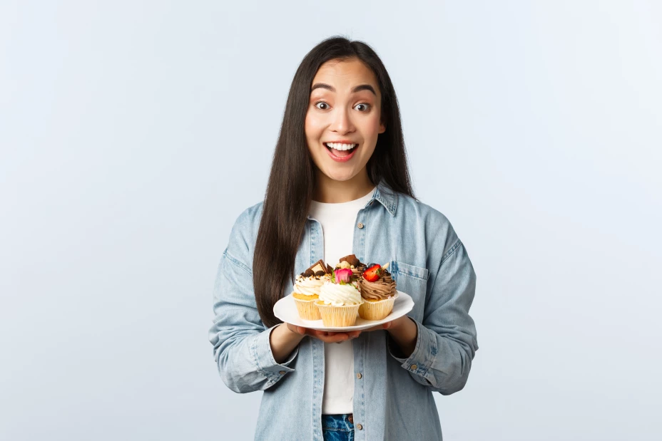 Social distancing lifestyle, covid-19 pandemic, celebrating holidays during coronavirus concept. Excited asian girl holding plate with tasty cupcakes, smiling broadly, invite visit cafe with muffins