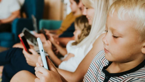 Niño pequeño usando un teléfono móvil mientras adultos y otros menores utilizan dispositivos electrónicos en el mismo espacio.