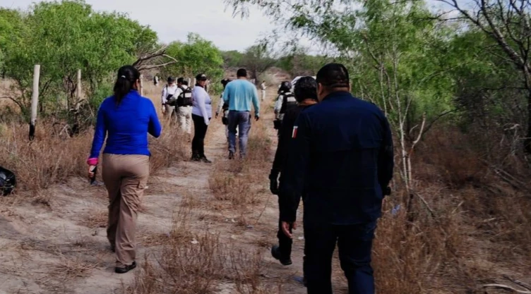 Fotografía de una brigada de búsqueda en un terreno árido de Tamaulipas. Se observa a un grupo de personas, integrantes del colectivo ‘Amor por los Desaparecidos’ y elementos de seguridad, caminando de espaldas por un sendero flanqueado por maleza seca y arbustos verdes. Al frente, una mujer con playera azul y pantalón café encabeza el grupo, mientras que otros visten uniformes tácticos o ropa de campo. El ambiente es soleado y el terreno es de tierra suelta, típico de las zonas de búsqueda de restos en el norte de México.