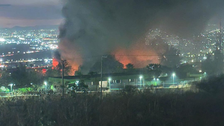 Fotografía nocturna del incendio de gran magnitud en una chatarrera de El Salto, Jalisco. En el centro, una intensa llamarada naranja ilumina una densa columna de humo negro que se eleva hacia el cielo oscuro. En primer plano se observa una barda perimetral iluminada y vegetación seca, mientras que al fondo se aprecian las luces de la ciudad bajo un cielo nocturno.
