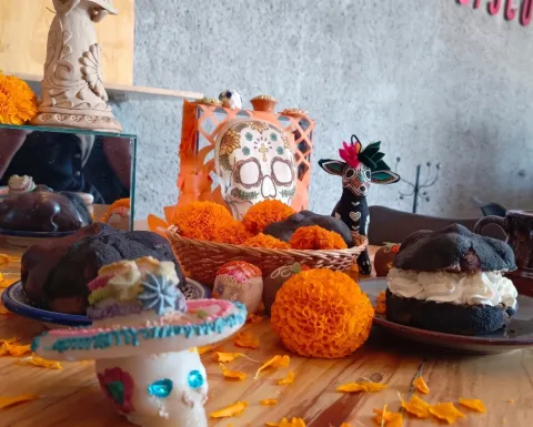 Mesa de madera decorada con piezas de pan de muerto negro, una rellena de crema chantilly blanca y otra con cubierta de chocolate, rodeadas de flores de cempasúchil, calaveritas de azúcar y una taza de barro.