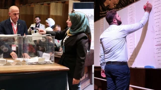 una mujer siria, de pie, deposita su voto en una urna electoral transparente dentro de un colegio electoral. su rostro muestra una expresión seria, reflejando la incertidumbre de las elecciones en la siria post-assad