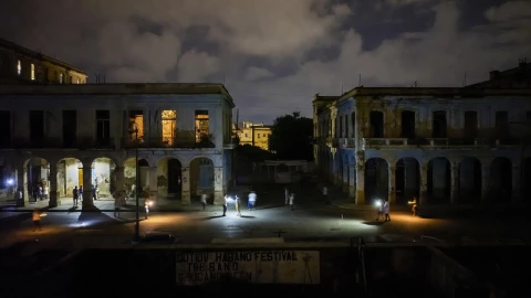 Fotografía periodística realista en orientación horizontal de La Habana durante un apagón nocturno, con calles parcialmente oscuras, edificios deteriorados apenas iluminados por luces aisladas, personas caminando con linternas o celulares, ambiente tenso y silencioso. En primer plano, un cartel apagado o deteriorado relacionado con el Festival del Habano, insinuando cancelación sin mostrar texto legible. Cielo nublado, iluminación tenue, estilo documental, alta resolución, sin texto, sin marcas de agua.