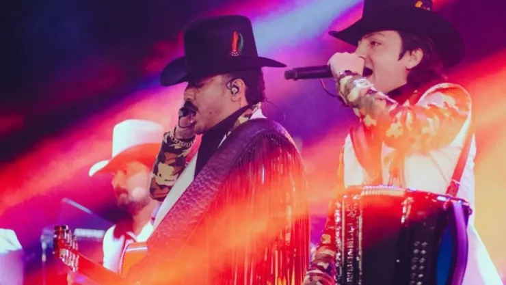 Dos hombres con un micrófono y cantando, con vestimenta grupera e instrumentos musicales