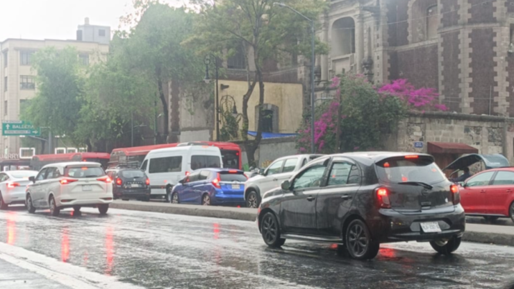 Esta es una toma a nivel de los ojos de un día de lluvia y nublado en la Ciudad de México, donde varios autos y camionetas se encuentran detenidos por el tráfico. La imagen está tomada sobre una calle donde se pueden observar el reflejo de las luces de los autos y el resto de la luz del día en el pavimento mojado, además de los autos en el tráfico de un día lluvioso. También hay varios árboles en la acera derecha, con una pared de cemento detrás de ellos que tiene algunas plantas moradas que caen en cascada sobre ella. Detrás de los árboles, hay un edificio de color melocotón con ventanas visibles, un muro que rodea el edificio de color melocotón y que parece una fortaleza, de color grisáceo y desgastado con el tiempo, y un edificio blanco que aparece en la parte izquierda. Además, en el lado izquierdo de la imagen hay un cartel de color blanco con letras y números en verde que dice “VALNERA”. En primer plano, en el lado izquierdo de la imagen, hay una minivan blanca con las luces traseras encendidas y detrás, un coche negro que parece un Nissan Versa, una minivan azul, una minivan blanca que parece una furgoneta, y un autobús rojo. El tráfico se detiene y la luz del día se desvanece de los autos en primer plano mientras se mueven hacia el fondo.