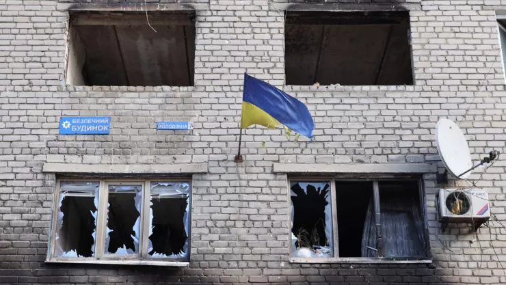 Edificio con ventanas estrelladas, así como fachada destruida, como si se hubiera incendiado. En el centro de la imagen aparece una bandera de Ucrania.