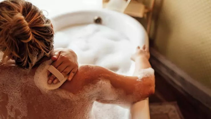 Una mujer dentro de una bañera con espuma mientras sostiene la esponja en su espalda