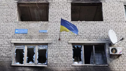 Edificio con ventanas estrelladas, así como fachada destruida, como si se hubiera incendiado. En el centro de la imagen aparece una bandera de Ucrania.