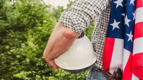 Un hombre sostiene un casco de obrero y una bandera de Estados Unidos.