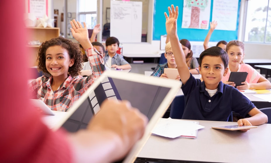 Un grupo de niños levantan la mano ante la maestra en un aula.
